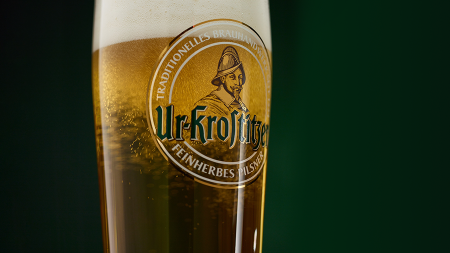 Ein Bierglas der Marke Ur-Krostitzer mit Bier gefüllt vor grünem Hintergrund