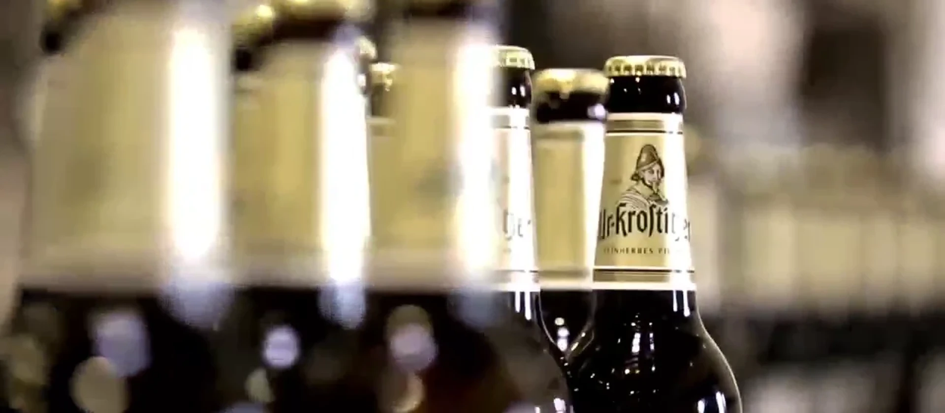 Mehrere braune Ur-Krostitzer Bierflaschen mit goldfarbenem Etikett und Kronkorken stehen in einer Reihe. Hintergrund unscharf.