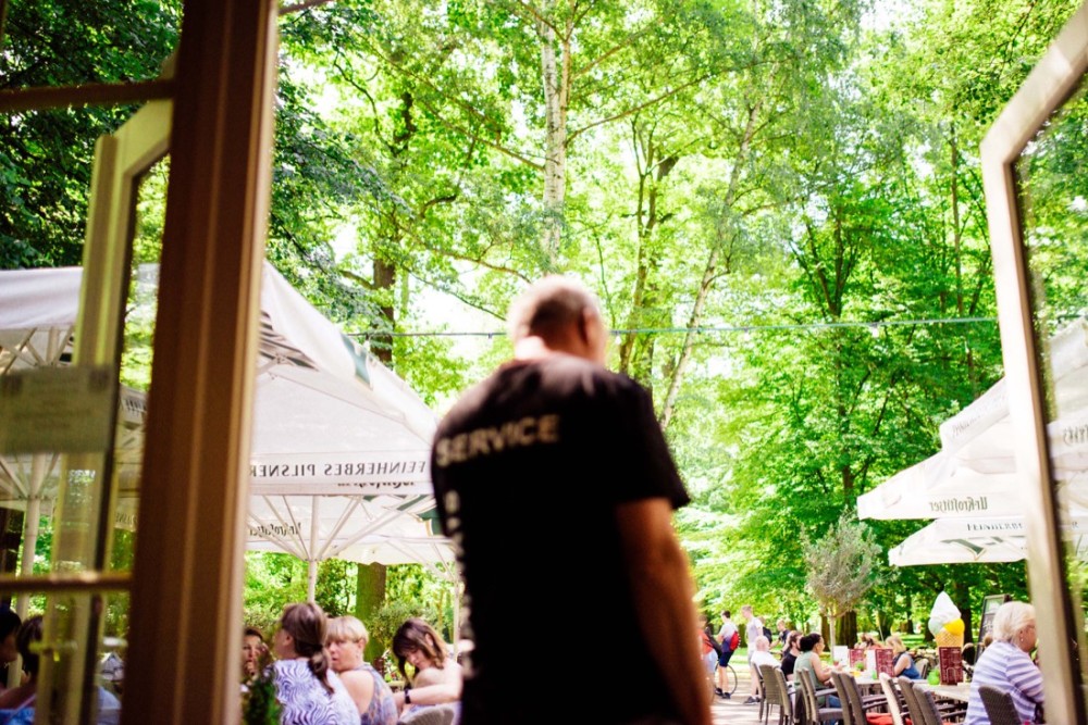 Ein Kellner mit schwarzer Kleidung geht in einem belebten Außenbereich eines Cafés zwischen Tischen hindurch.