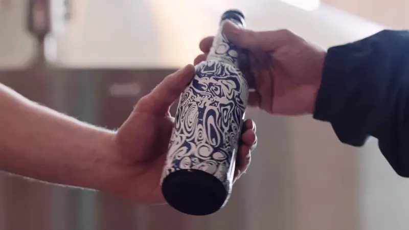 Zwei Hände halten eine Flasche mit blau-weißem, abstrakt gemustertem Etikett. Der Hintergrund ist unscharf und hell.