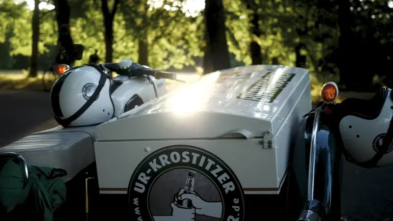 Weißes Motorrad mit Beiwagen und Ur-Krostitzer-Logo steht im Sonnenschein in einem Waldstück.