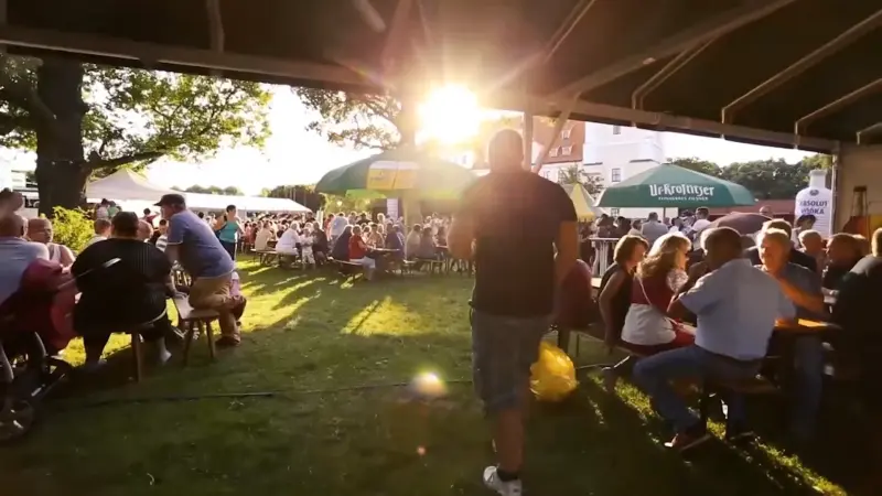 Menschen sitzen an Biertischen unter Ur-Krostitzer Sonnenschirmen bei Abendsonne. Im Hintergrund Zelte und Bäume.