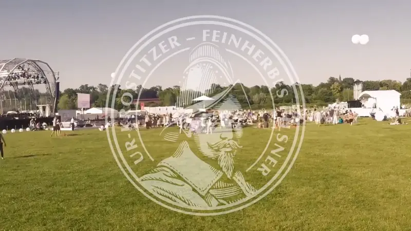 Weitläufige Wiese mit Festivalbühne und Besuchern bei Sonnenschein. Im Vordergrund ein transparentes Ur-Krostitzer-Logo.