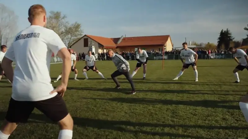 Fußballspieler in weißen Trikots wärmen sich auf einem Rasenplatz auf. Im Hintergrund stehen Zuschauer und Gebäude.