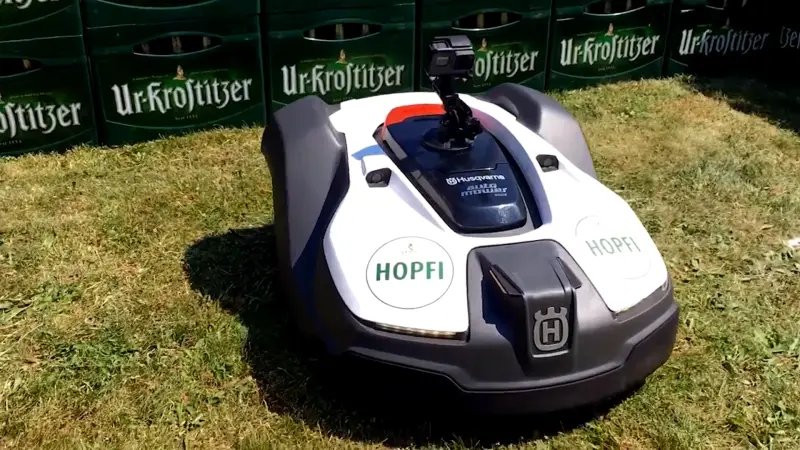 Weißer Mähroboter mit Kamera steht auf Rasen vor gestapelten grünen Ur-Krostitzer Bier-Getränkekisten.