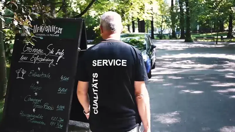 Mann im schwarzen T-Shirt mit der Aufschrift Service steht vor einer Ur-Krostitzer-Getränketafel in einem Park mit Bäumen.