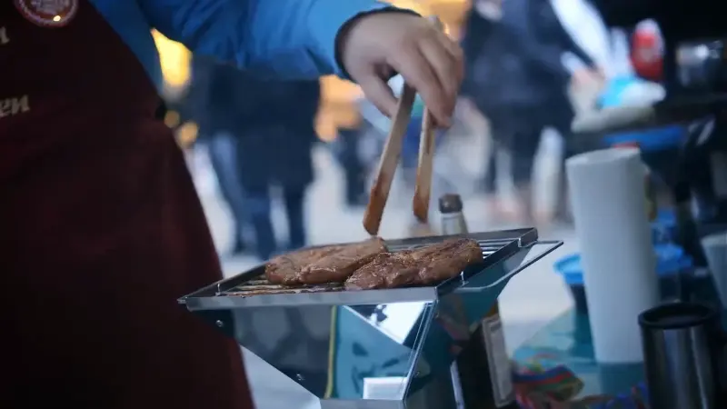 Hand wendet Fleischstücke mit einer Zange auf einem kleinen Grill. Der Hintergrund ist unscharf mit Personen.