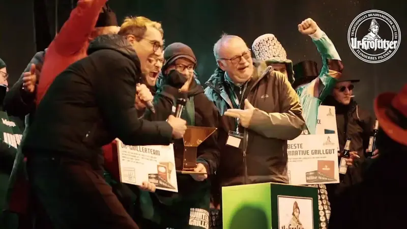 Gruppe von Personen feiert auf einer Bühne mit Urkunden und Trophäe. Einige halten Ur-Krostitzer Bierflaschen in der Hand.