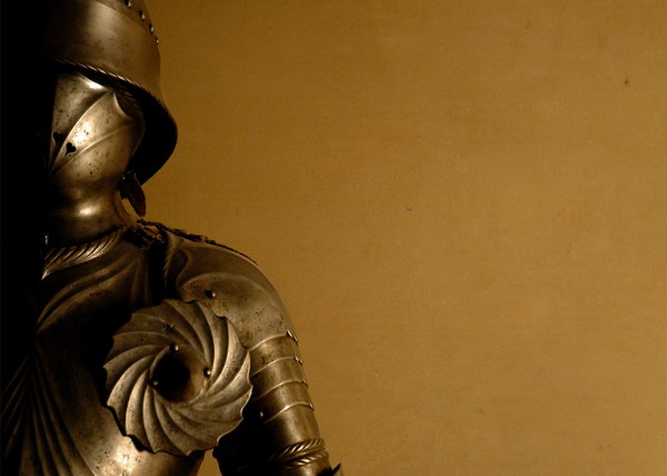 Ein mittelalterlicher Ritterhelm ist neben einem Teil der Rüstung zu sehen. Die Beleuchtung hebt metallische Details hervor.