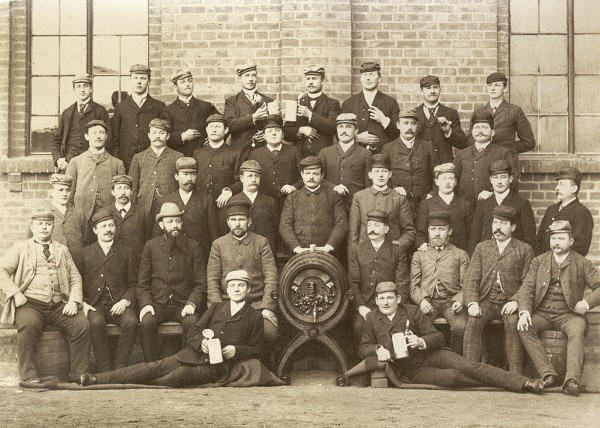 Eine Gruppe von Männern posiert in historischen Anzügen vor einem Gebäude. Sie tragen Mützen und halten teilweise Flaschen.