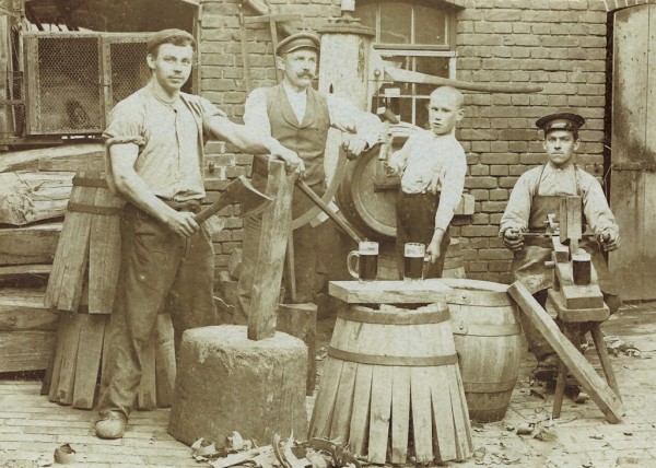 Vier Männer in traditioneller Arbeitskleidung posieren mit Werkzeugen und Holzfässern.