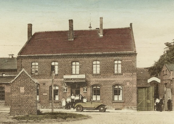 Ein historisches Foto zeigt ein Backsteinhaus mit Personen und einem Auto davor.