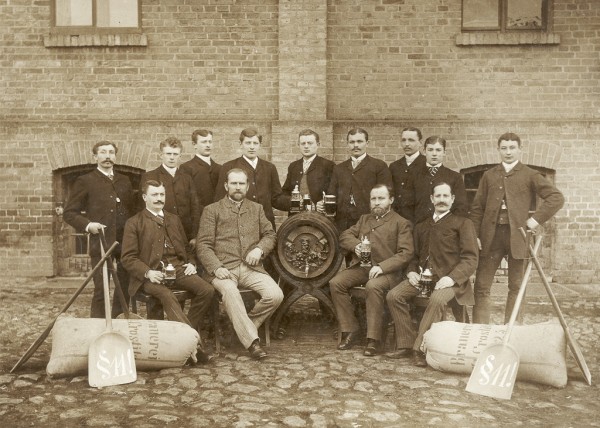 Zwölf Männer posieren mit Pokalen und Schaufeln vor einer Backsteinmauer.