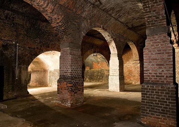 Eine alte Ziegelgewölbehalle mit Bögen und Säulen ist beleuchtet.