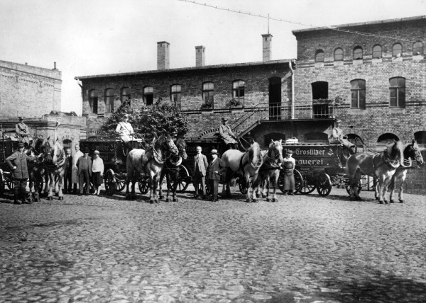 Vor einem historischen Gebäude stehen mehrere Pferdekutschen mit Arbeitern und Pferden.