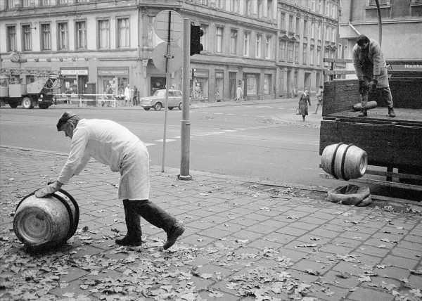 Ein Mann in einem weißen Kittel rollt ein großes Fass auf einer Straße. Ein anderer Mann hebt ein Fass auf einen LKW.