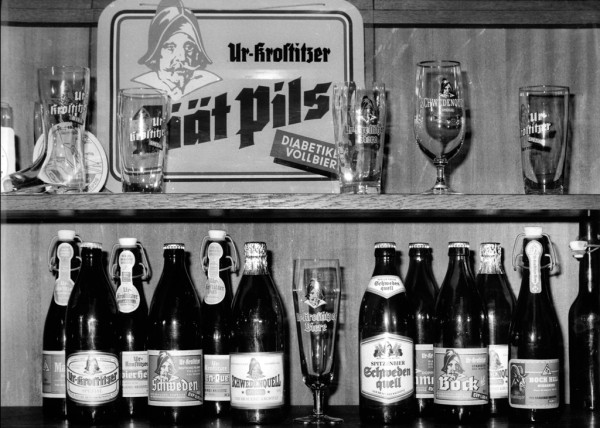 Verschiedene Bierflaschen und -gläser sind auf Holzregalen ausgestellt. Ein Werbeschild befindet sich im Hintergrund.