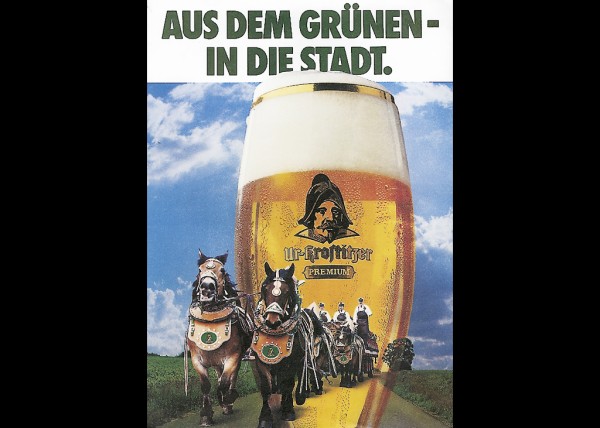 Zwei Pferde ziehen eine Kutsche auf einer Straße, daneben ein großes Bierglas.