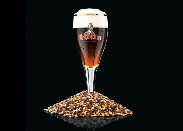 Ein Glas dunkles Bier steht auf einem Haufen Gerstenkörner vor schwarzem Hintergrund.