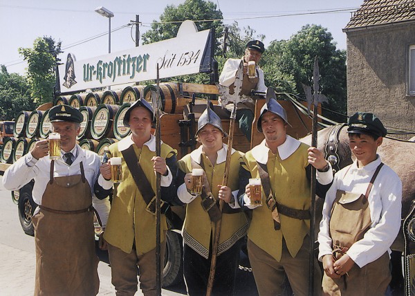 Sechs Personen in traditioneller Kleidung halten Bierkrüge vor einem Bierwagen.