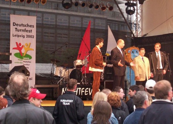 Mehrere Personen stehen auf einer Bühne bei einer Veranstaltung im Freien. Ein blaues Maskottchen ist auch dabei.
