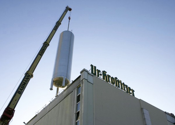Ein Kran hebt einen großen zylindrischen Tank auf ein Fabrikgebäude.