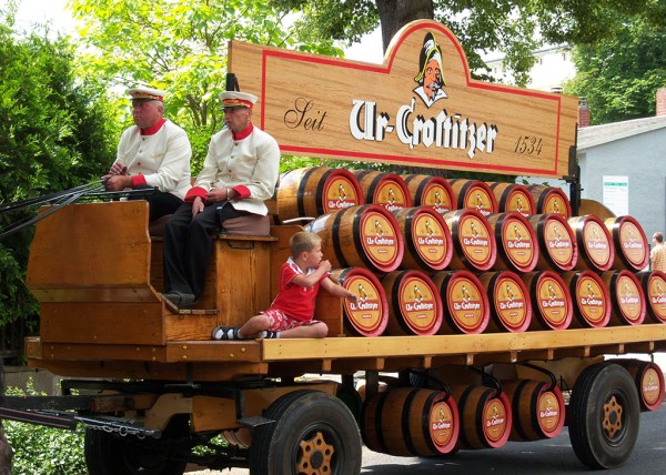 Zwei Männer in traditioneller Kleidung sitzen auf einem alten Holzwagen, beladen mit vielen Fässern.