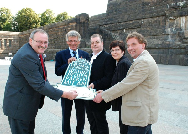 Fünf Personen übergeben ein Plakat vor einer großen Steintreppe.