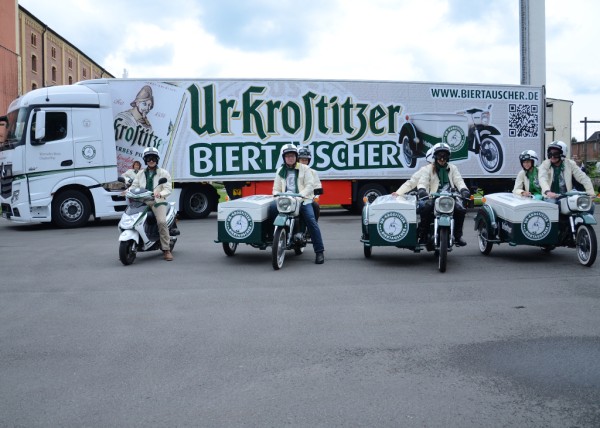 Vier Personen fahren auf weißen Motorrollern vor einem geparkten LKW. Alle tragen weiße Helme und Jacken.