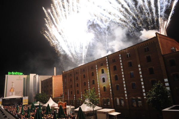 Feuerwerk erleuchtet den Nachthimmel über einem belebten Marktplatz neben alten Fabrikgebäuden.