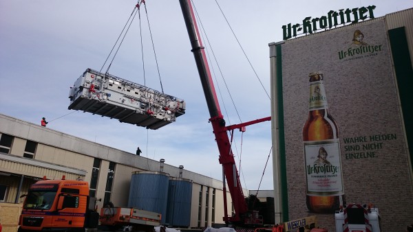 Ein großer Kran hebt eine industrielle Maschine neben einem Gebäude mit einem Bierwerbeplakat.