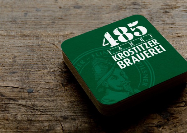 Ein grüner Bierdeckel mit einer historischen Illustration liegt auf einer Holzoberfläche.