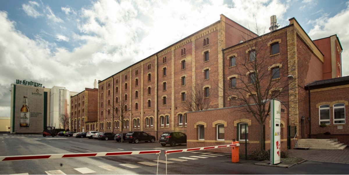 Lange Ziegelgebäude mit vielen Fenstern unter bewölktem Himmel, davor eine Schranke und Autos.