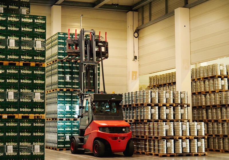Ein roter Gabelstapler hebt in einer Lagerhalle eine Last aus mehreren Bierfässern.