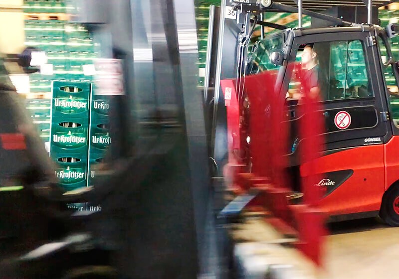 Ein Gabelstapler bewegt sich in einer Lagerhalle vorbei an einer Palette mit Getränkekisten. Der Fahrer ist sichtbar und konzentriert.
