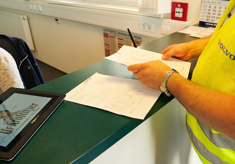 Ein Mann in einer Warnweste übergibt Dokumente an eine Person an einem Schreibtisch.