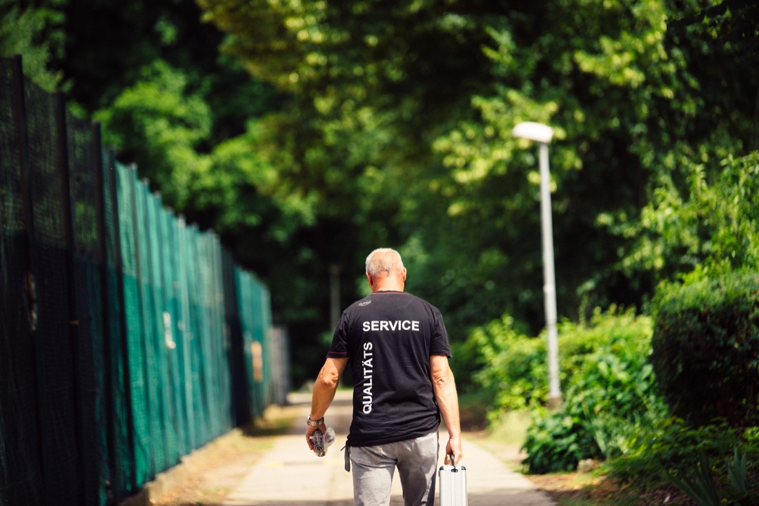 Ein Mann geht an einem sonnigen Tag auf einem Gehweg neben einem grünen Zaun. Er trägt ein schwarzes T-Shirt und hält eine Tasche.