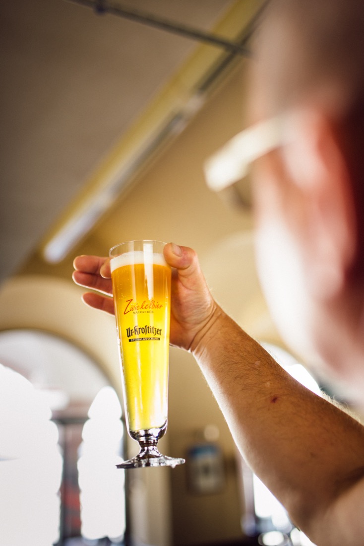 Eine Person hält ein volles Bierglas gegen das Licht. Das Glas steht auf einem eleganten Fuß.
