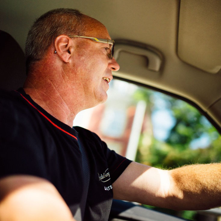 Ein älterer Mann fährt lächelnd ein Auto an einem sonnigen Tag.