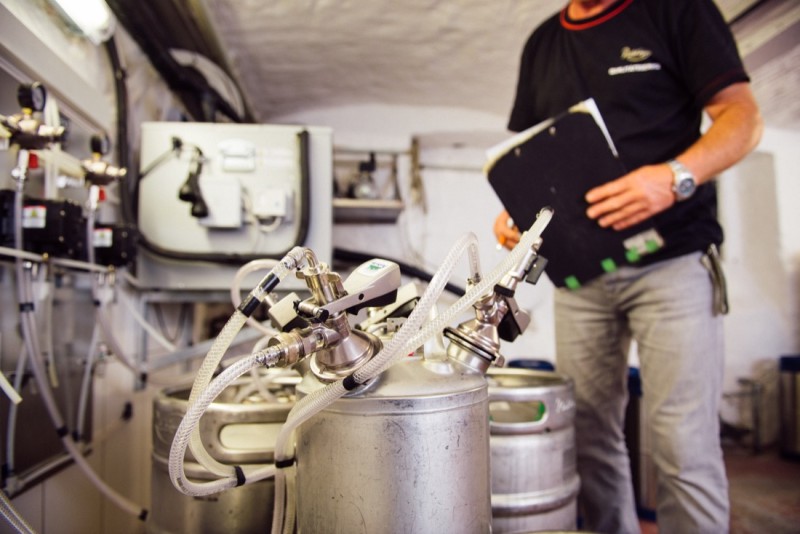 Ein Mann steht neben Metallfässern und Maschinen in einer Brauerei. Er hält ein Klemmbrett.