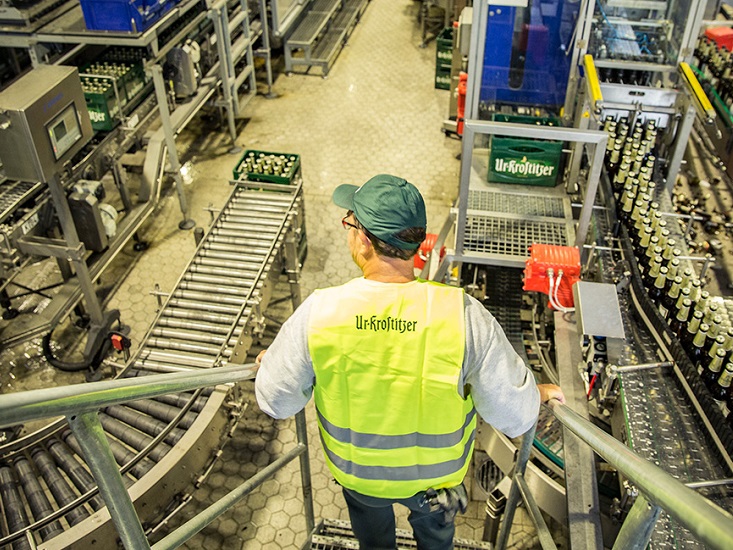 Ein Mann in gelber Warnweste beobachtet eine Flaschenabfüllanlage von einer erhöhten Plattform.