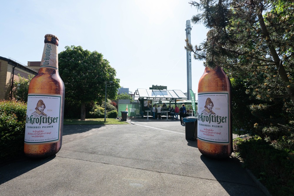 Zwei große Bierflaschen flankieren einen Weg zu einem Imbissstand auf einem sonnigen Platz.
