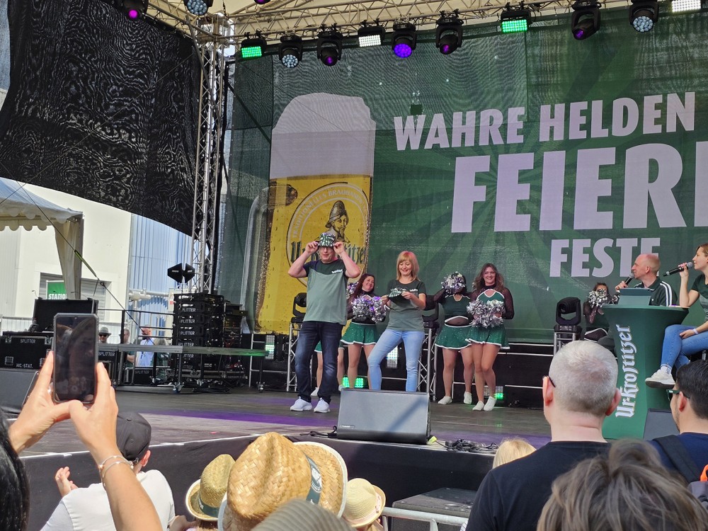 Mehrere Personen stehen auf einer Bühne bei einem öffentlichen Event. Einige tanzen, während andere zuschauen.