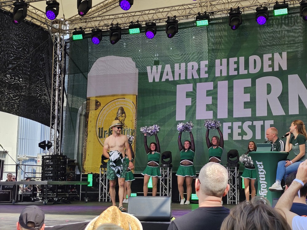 Eine Bühnenszene bei einem Festival mit Cheerleadern, einem Moderator und einem kostümierten Mann.