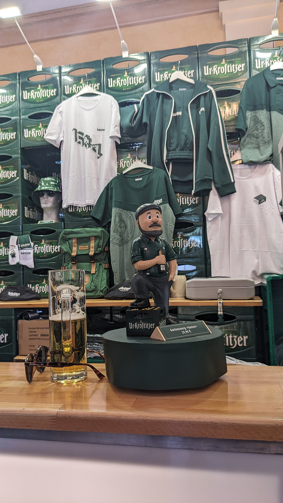 Ein Bierglas steht vor einer modellierten Figur in einer Ladenumgebung mit thematischen Textilien im Hintergrund.