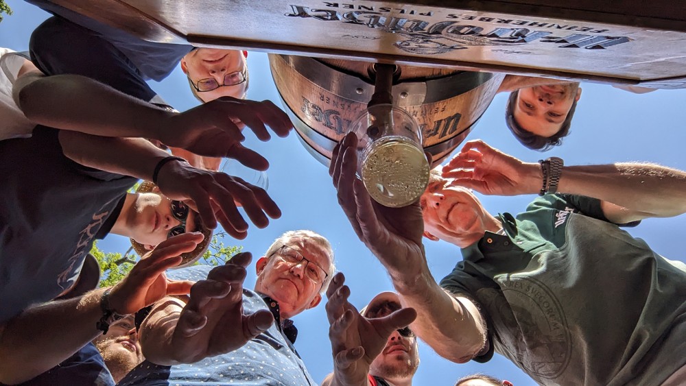 Mehrere Personen greifen nach einem Glas Bier, das von unten fotografiert wird.