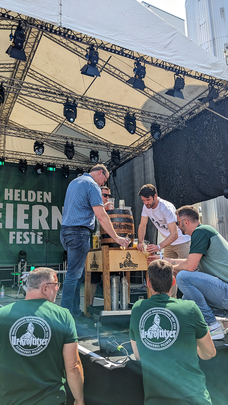 Männer zapfen Bier an einem Holzfass auf einer Bühne, umgeben von Zuschauern.