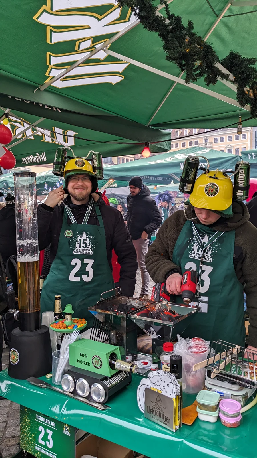 Zwei Personen verkaufen Lebensmittel an einem Marktstand, eine davon trägt einen Hut mit Getränkedosen.