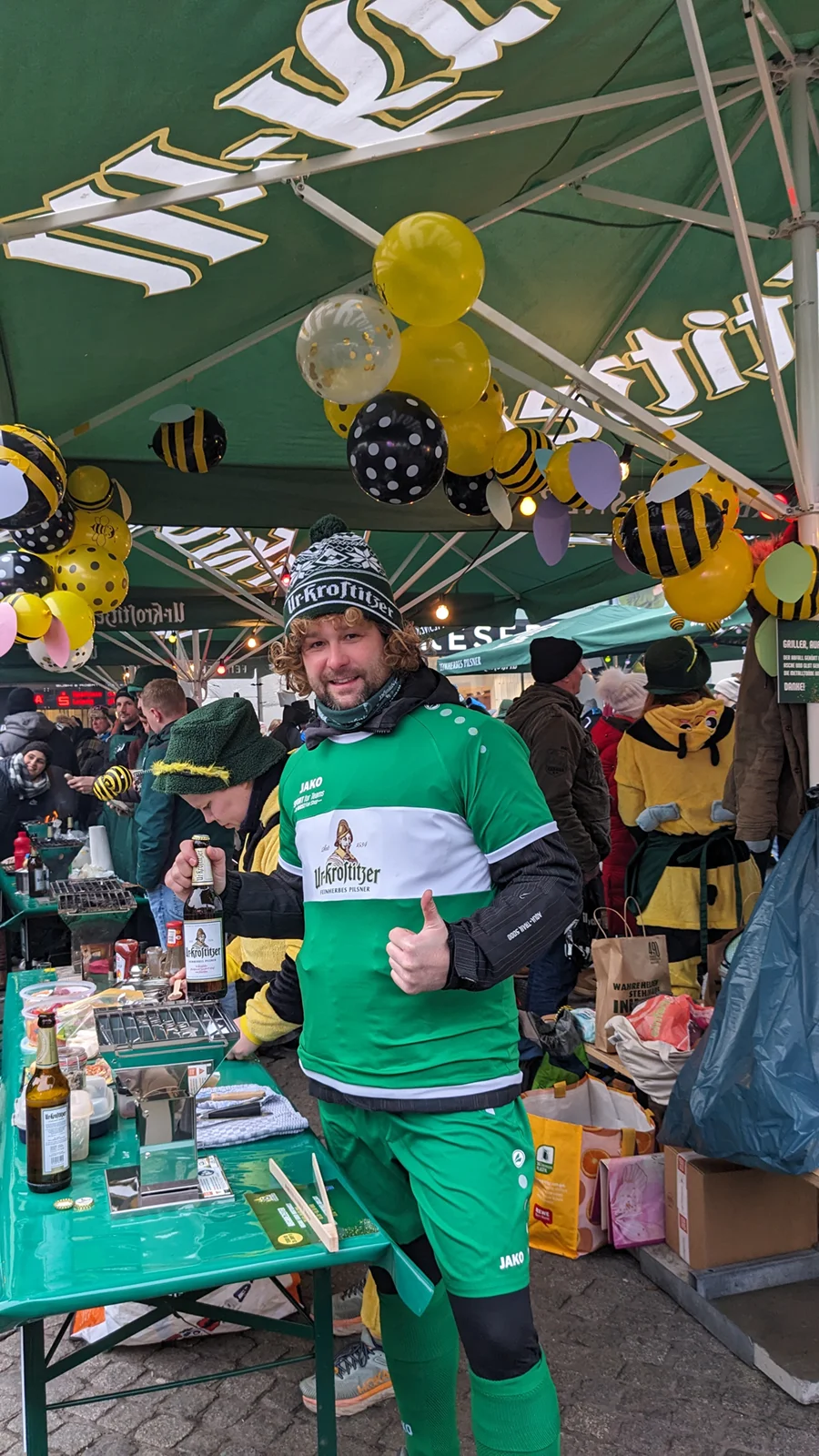 Ein Mann in grünem Sportoutfit zeigt Daumen hoch unter einem Partyzelt mit gelb-schwarzen Ballons.