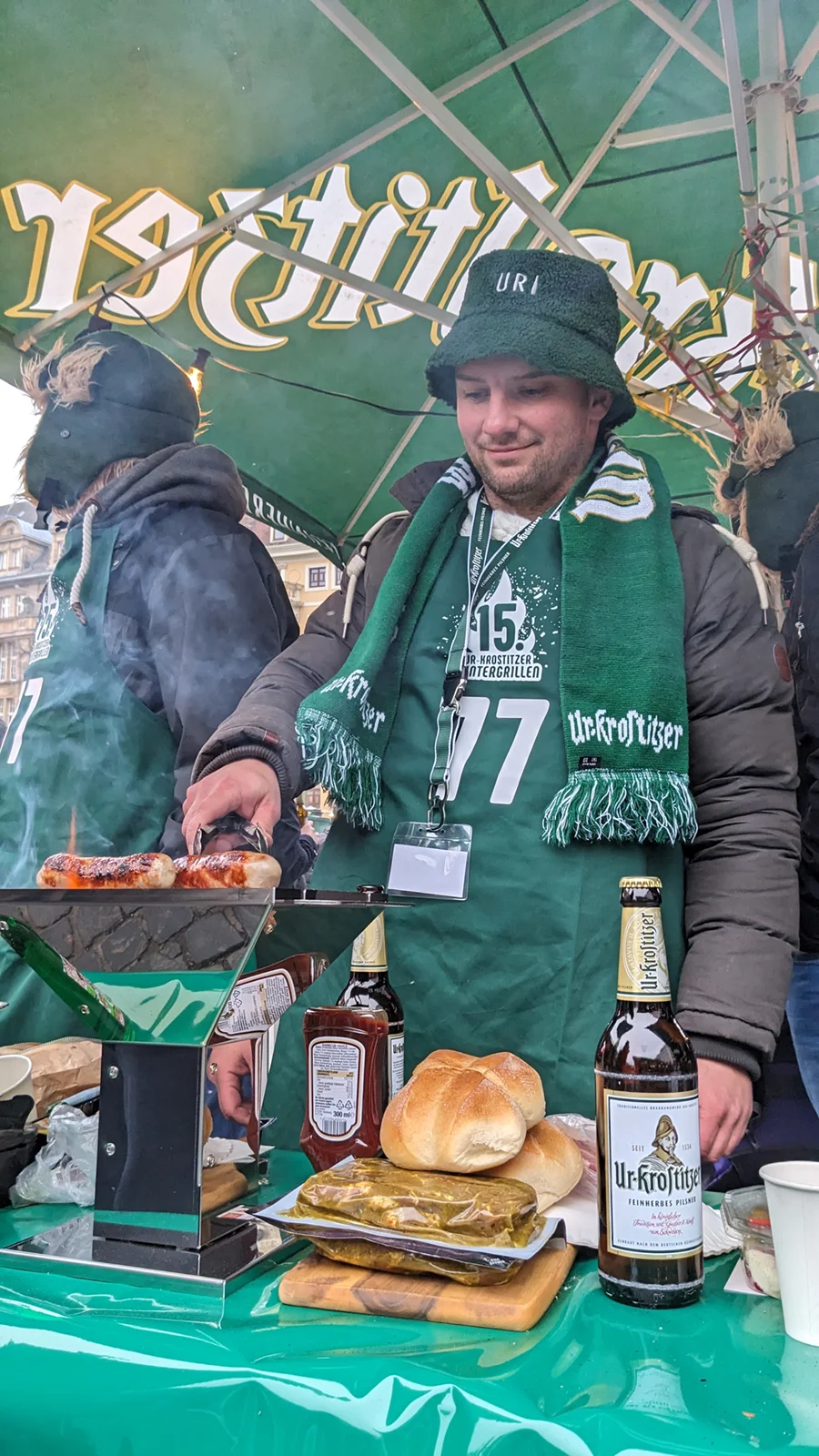 Ein Mann in grüner Kleidung brät Würstchen auf einem Marktstand. Auf dem Tisch stehen Bier und Brötchen.